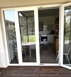 Ein heller Wohnraum mit offenen Fenstern und Blick auf einen grünen Garten. Im Innenraum sind eine Küche und ein Essbereich sichtbar.