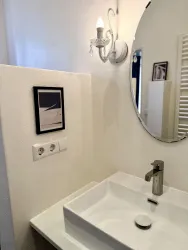 Ein modernes Bad mit einem weißen Waschbecken und einem runden Spiegel. An der Wand hängt ein Bild in einem schwarzen Rahmen.