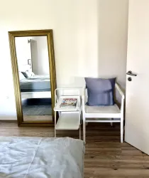Ein helles Schlafzimmer mit einem großen Spiegel und einem weißen Stuhl neben einem kleinen Tisch. Auf dem Tisch liegen Zeitschriften, und es gibt einen blauen Kissen auf dem Stuhl.