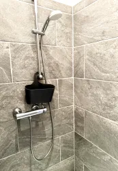 Eine moderne Dusche mit grauen Fliesen und einem Handbrausehalter. Der Duschkopf ist an der Wand montiert, und ein schwarzes Ablagefach ist sichtbar.