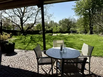 Ein gemütlicher Gartentisch mit Stühlen steht im Freien. Der Blick zeigt eine grüne Wiese und blühende Bäume im Hintergrund.