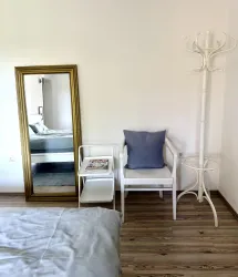 Ein modernes Schlafzimmer mit einem großen Spiegel und einem weißen Stuhl. Neben dem Stuhl steht ein Kleidungshalter und ein kleiner Tisch mit Zeitschriften.