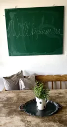 Ein einladender Tisch mit einem rustikalen Holztisch und einem kleinen Topf mit Kräutern. Im Hintergrund hängt eine grüne Tafel mit dem Wort "Willkommen".