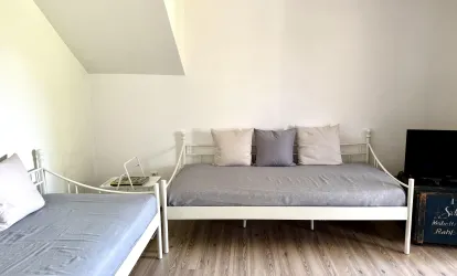 Ein einfaches Zimmer mit zwei Betten und neutralen Farbtonen. Es gibt Kissen und einen kleinen Fernseher in der Ecke.