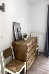 Ein heller Raum mit einem Holzschrank und einem gemütlichen Stuhl. An der Wand hängt ein schwarz-weißes Bild, und auf dem Schrank steht eine kleine Pflanze in einem weißen Topf.