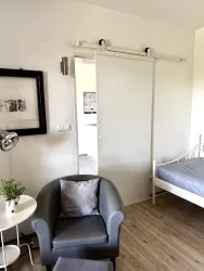 Ein modernes Zimmer mit einem grauen Sessel, einem kleinen Tisch und einem Bett mit grauer Bettwäsche. Eine Schiebetür führt zu einem weiteren Raum, der teilweise sichtbar ist.