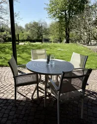 Ein runder Tisch mit vier Stühlen steht auf einem gepflasterten Boden. Im Hintergrund ist eine grüne Wiese und Bäume im Sonnenschein zu sehen.