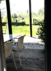 Ein schöner Blick von einem Innenraum auf eine Terrasse mit einem Tisch und Stühlen. Im Hintergrund sieht man eine grüne Wiese und Bäume.