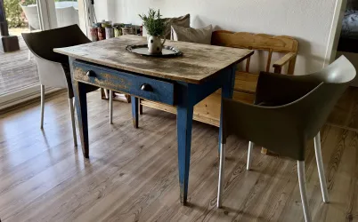 Ein Holzttisch mit blauen Beinen steht in einem gemütlichen Raum. Um den Tisch herum sind zwei moderne Stühle platziert.
