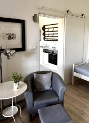 Ein modernes Zimmer mit einem bequemen Sessel und einem kleinen Tisch. Im Hintergrund ist eine offene Küche sichtbar.