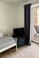 Ein einfaches Zimmer mit einem Bett, einem Fernseher und einem alten Holzkasten. Durch die große Glas-Tür gelangt Licht und man sieht einen Außenbereich.