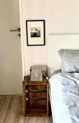Ein gemütliches Schlafzimmer mit einem Bett und einem Holzregal. An der Wand hängt ein gerahmtes Bild, und auf dem Regal steht eine Glaslampe.