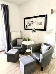 Ein gemütlicher Raum mit zwei grauen Sessel und einem kleinen Tisch. An der Wand hängt ein gerahmtes Kunstwerk, und es gibt eine Stehlampe sowie Hocker.