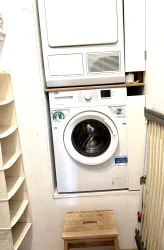 Eine Waschmaschine und ein Wäschetrockner stehen übereinander in einem kleinen Raum. Daneben befindet sich eine Holzbank und ein Regal.