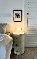 Ein minimalistisches Schlafzimmer mit einem runden Nachttisch und einer Lampe. An der Wand hängt ein gerahmtes Kunstwerk.