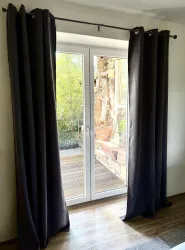 Ein heller Raum mit großen Fenstertüren, die von langen, dunklen Vorhängen umgeben sind. Im Hintergrund sind grüne Pflanzen und ein Holzboden sichtbar.