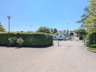 Ein gepflasterter Weg führt zu einem Bereich mit Bäumen und Büschen. Im Hintergrund ist ein weißer Wohnwagen und einige Parkplätze zu sehen.