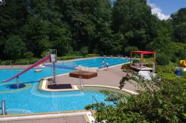 Ein Schwimmbad mit einer Rutsche und einer großzügigen Liegewiese. Rundherum sind Bäume und eine entspannte Atmosphäre.
