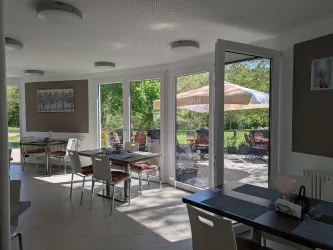 Ein helles und einladendes Café mit großen Fenstern und Blick auf den Außenbereich. Tische und Stühle sind ordentlich angeordnet, und Sonnenschirme stehen im Freien.