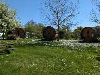 Drei gemütliche Holzhütten stehen in einer blühenden Wiese. Im Hintergrund sind Bäume und blauer Himmel zu sehen.