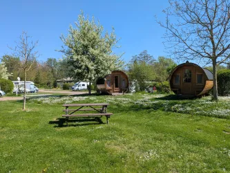 Ein ruhiger Campingplatz mit zwei gemütlichen Holzhütten und einem Picknicktisch. Die Wiese ist grün und blühend, umgeben von Bäumen und klarem Himmel.
