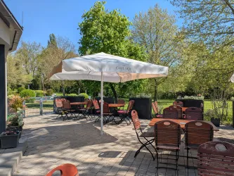 Ein einladender Außenbereich mit Tischen und Stühlen unter einem großen Sonnenschirm. Umgeben von Bäumen und grünem Gras.