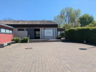 Ein modernes Gebäude mit einer gepflegten Eingangsfläche. Auf der rechten Seite ist ein begrüntes Gelände sichtbar.