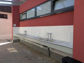 Eine schlichte Außenküche mit mehreren Spülen ist zu sehen. Die Wand ist rot und die Umgebung ist hell und gut beleuchtet.