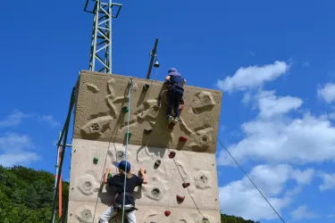 Zwei Kinder klettern an einer Kletterwand unter einem klaren, blauen Himmel. Die Wand ist mit bunten Griffen ausgestattet und bietet eine spannende Herausforderung.