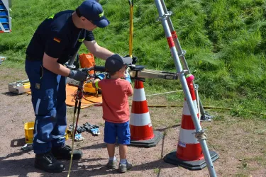 Ein Erwachsener arbeitet an einem technischen Gerät, während ein Kind beobachtet und lernt. Im Hintergrund sind Absperrkegel und Werkzeuge zu sehen.