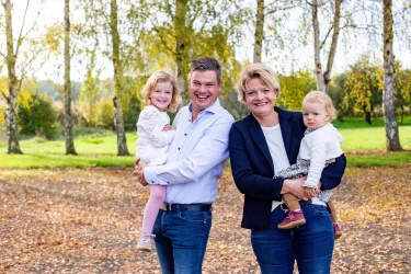 Eine glückliche Familie steht im Freien zwischen Bäumen. Sie halten zwei kleine Mädchen im Arm und lächeln freundlich.