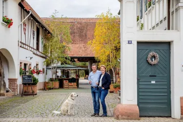 Ein idyllischer Innenhof mit zwei Personen und einem Hund. Umgeben von historischen Gebäuden und herbstlichen Bäumen.
