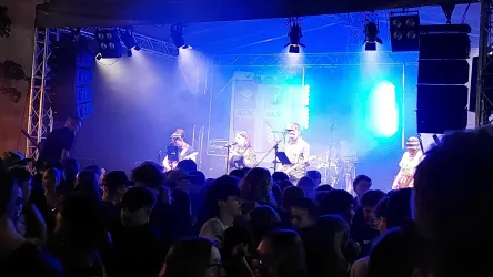 Eine Band spielt auf einer Bühne vor einem begeisterten Publikum. Die Bühne ist mit bunten Lichtern beleuchtet und es herrscht eine fröhliche Stimmung.