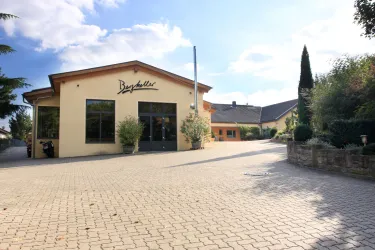 Ein modernes Gebäude mit der Aufschrift "Bayernstube" und einer gepflasterten Auffahrt. Die Umgebung ist grün und gepflegt, unter einem klaren blauen Himmel.