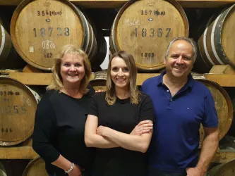 Drei Personen stehen vor traditionellen Holzfässern. Sie lächeln und wirken freundlich.