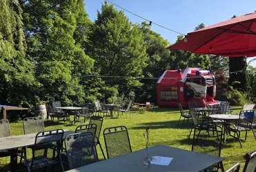 Ein gemütlicher Außenbereich mit Tischen und Stühlen auf einer Wiese. Im Hintergrund steht ein roter Verkaufswagen und Bäume spenden Schatten.