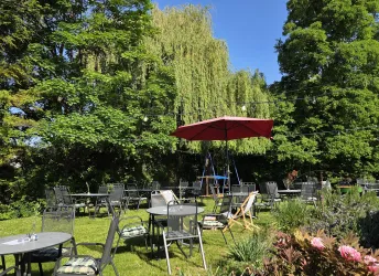 Ein schöner Garten mit Tischen und Stühlen unter einem Sonnenschirm. Umgeben von grünen Bäumen ist es ein einladender Ort zum Entspannen.