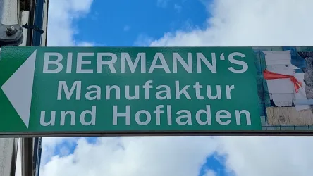 Ein Straßenschild zeigt auf die "Biermann's Manufaktur und Hofladen". Der Himmel ist blau mit einigen Wolken.