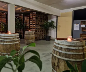 Ein gemütlicher Raum mit Holzfässern und Kerzenlicht. Im Hintergrund sind Regale mit Kisten und einige Pflanzen zu sehen.