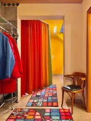 Ein farbenfroher Flur mit Vorhängen in Rot, Orange und Grün. Ein bunter Teppich und ein Stuhl verleihen dem Raum eine einladende Atmosphäre.