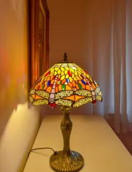 Eine bunte Tiffany-Lampe steht auf einem Tisch. Ihr Schirm besteht aus farbigen Glasscheiben und wirft warmes Licht in den Raum.