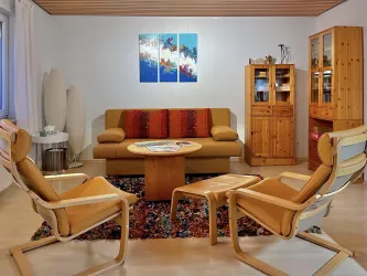 Ein gemütliches Wohnzimmer mit einer beigen Couch und zwei modernen Stühlen. An der Wand hängen Bilder, und Möbel aus Holz verleihen dem Raum einen warmen Charakter.