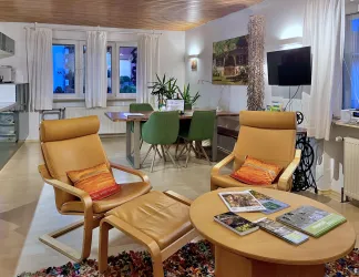 Ein gemütlicher Wohnraum mit modernen Möbeln, gepolsterten Sesseln und einem runden Tisch. Pflanzen und eine angenehme Beleuchtung schaffen eine einladende Atmosphäre.