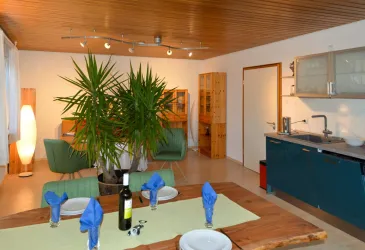 Ein modernes und einladendes Wohnzimmer mit einem großen Esstisch, grünen Stühlen und einer Pflanze. Die offene Küche ist gut ausgestattet und schafft eine gemütliche Atmosphäre.