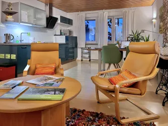 Ein modernes Wohnzimmer mit zwei bequemen Sesseln und einem runden Tisch. Die Küche ist gut ausgestattet und es gibt große Fenster, die für viel Tageslicht sorgen.