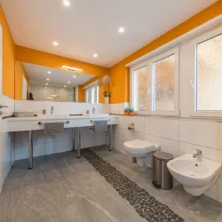 Ein modernes Badezimmer mit orangefarbenen Wänden und hellen Fliesen. Es gibt einen Doppelwaschtisch, eine Toilette und einen Bidet, sowie große Fenster für viel Tageslicht.