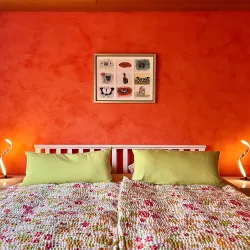 Ein gemütliches Schlafzimmer mit einem orangefarbenen Wanddesign. Das Bett hat bunte Decken und zwei Nachttischlampen.