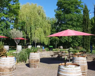 Ein einladender Außenbereich mit Holztischen und roten Sonnenschirmen. Üppige Bäume und fassartige Tischdekoration schaffen eine gemütliche Atmosphäre.