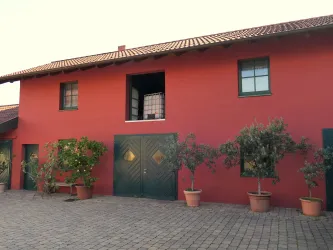 Ein rotes Gebäude mit einer grauen Tür und mehreren Fenstern. Vor dem Gebäude stehen Topfpflanzen und es gibt einen gepflasterten Bereich.
