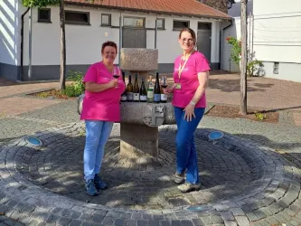 Zwei Frauen in pinken T-Shirts stehen an einem Brunnen und halten Weingläser. Hintergrund zeigt eine ruhige, gepflasterte Umgebung.